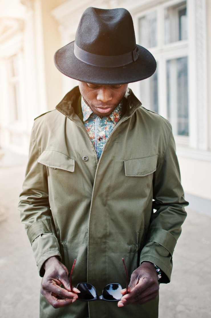 Gratis Foto close-up mode portret van zwarte afro-amerikaanse man op groene jas mantel en zwarte hoed met zonnebril verblijf achtergrond geel herenhuis verticale foto