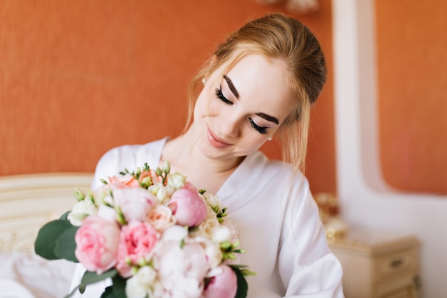 Close-up portret gelukkige bruid in witte badjas in de ochtend. Ze kijkt naar boeket bloemen in handen en lacht