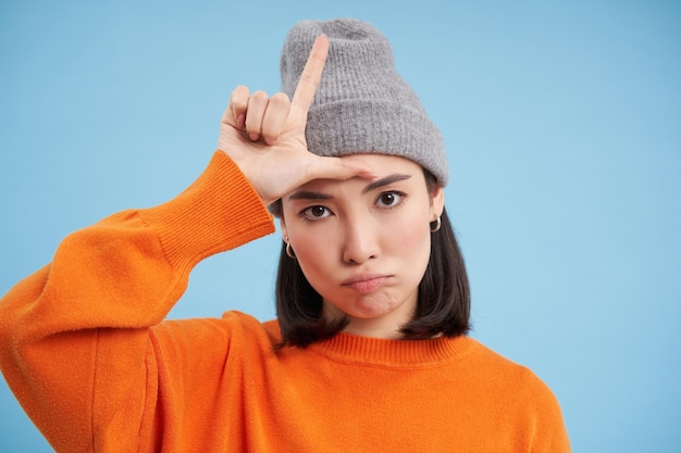 Gratis foto close-up portret van aziatische studente in beanie toont l letter verliezer teken op voorhoofd staat voorover