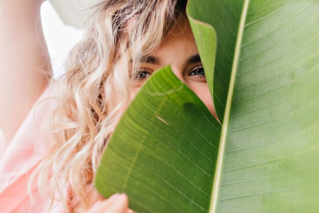 Close-up portret van blauwogige meisje speels poseren met plant. charmante verlegen blonde dame gezicht achter groen blad verbergen.