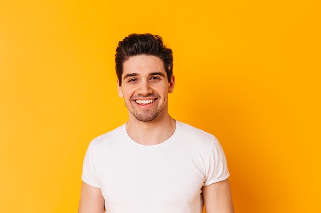 Close-up portret van bruinogige man in wit t-shirt. Guy glimlacht op oranje ruimte.