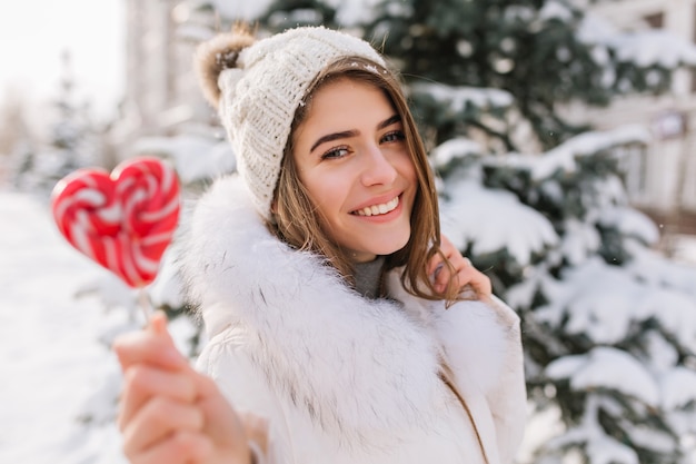 Close-up portret van charmante dame in witte jas met zoete lolly. Buitenfoto van zalige blonde vrouw in gebreide muts poseren naast boom in winterdag met rode kandijsuiker.