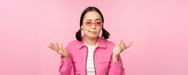 Gratis foto close-up portret van een aziatisch meisje dat verward haar schouders ophaalt en naar de camera kijkt met een zonnebril op die over een roze achtergrond staat