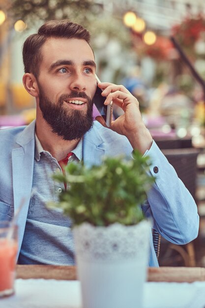 Close-up portret van een glimlachende modieuze bebaarde zakenman met een stijlvol kapsel, sprekend via de telefoon, drinkt een glas sap, zittend in een café buiten.