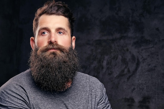 Close-up portret van een hipster met volle baard en stijlvol kapsel, gekleed in een grijs t-shirt, staat in een studio op een donkere achtergrond.