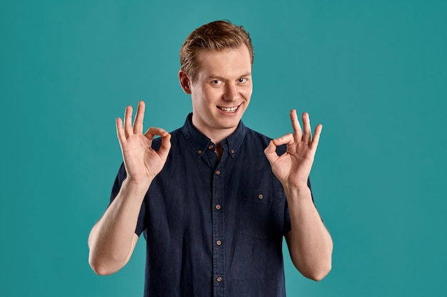 Close-up portret van een jonge atletische gember man in een stijlvol marine t-shirt glimlachend, ok teken tonen en kijken naar de camera terwijl poseren op blauwe studio achtergrond. menselijke gezichtsuitdrukkingen. eerlijk