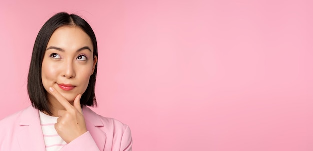 Close-up portret van een jonge Aziatische zakenvrouw die nadenkend glimlacht en naar de linkerbovenhoek kijkt die over een roze achtergrond staat