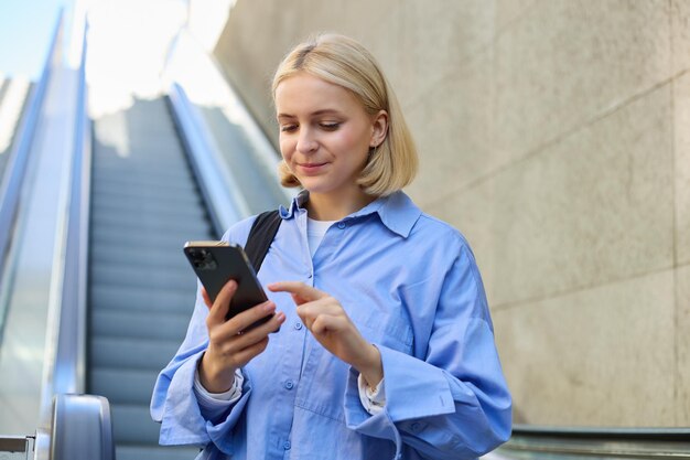 Gratis foto close-up portret van een jonge blonde vrouw in het stadscentrum die bij de roltrap staat en haar mobiele telefoon controleert