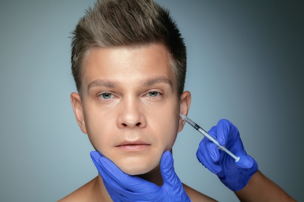 Close-up portret van een jonge man geïsoleerd op een grijze muur. Operatieprocedure vullen. Concept van de gezondheid en schoonheid van mannen, cosmetologie, zelfzorg, lichaams- en huidverzorging. Anti-veroudering.