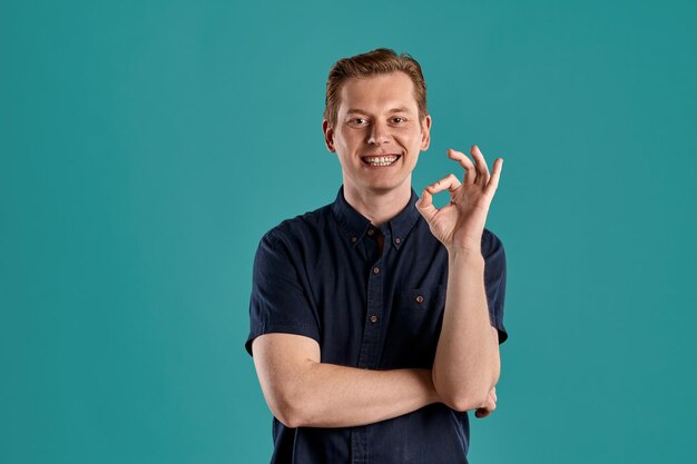 Close-up portret van een jonge statige gember kerel in een stijlvol marine t-shirt glimlachend, ok teken tonen en kijken naar de camera terwijl poseren op blauwe studio achtergrond. Menselijke gezichtsuitdrukkingen. Oprechte