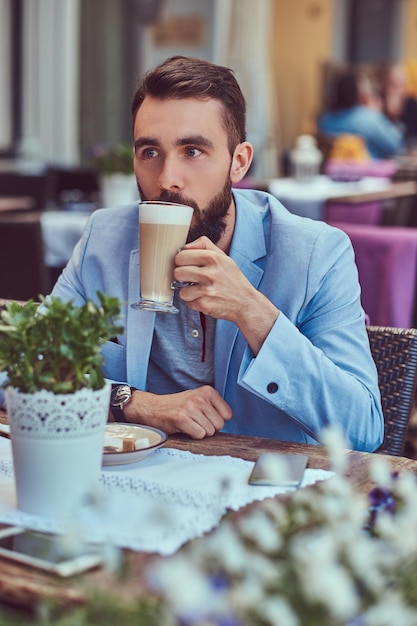 Close-up portret van een modieuze bebaarde man met een stijlvol kapsel, drinkt een glas cappuccino, zittend in een café buiten.