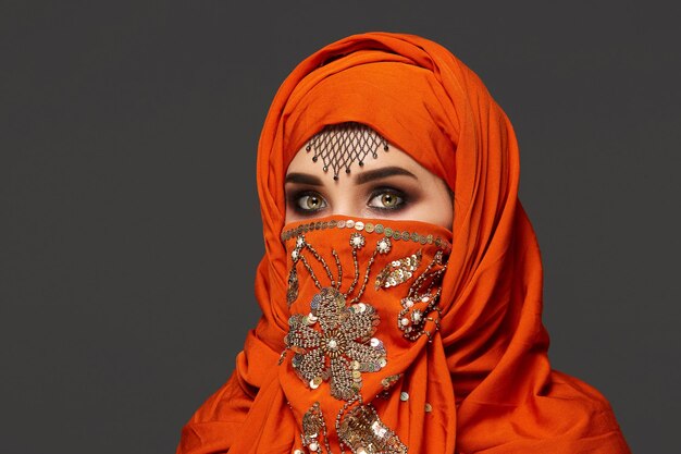 Close-up portret van een mooie jonge vrouw met rokerige ogen en fijne sieraden op het voorhoofd, gekleed in de terracotta hijab versierd met pailletten. Ze poseert en kijkt naar de camera op een donkere ba