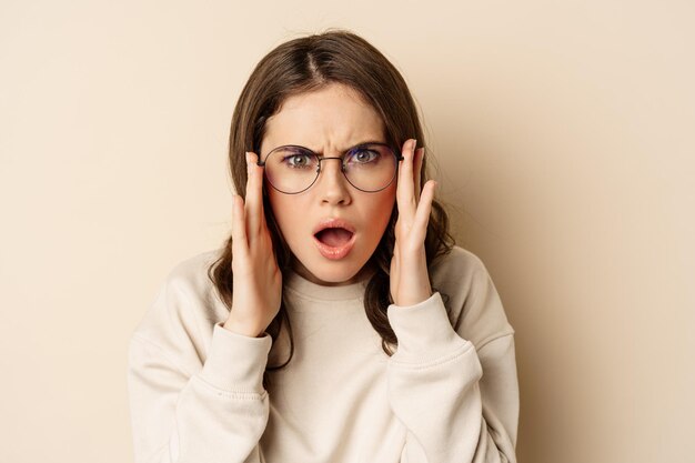 Close-up portret van een vrouw met een bril die er verward en gefrustreerd uitziet, kan iets vreemds niet begrijpen, staande over een beige achtergrond.