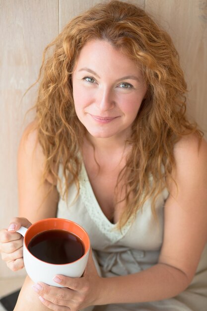 Close-up portret van een vrouw van middelbare leeftijd die hete zwarte thee drinkt. Gelukkige dame met rood haar die lacht voor de carema bij haar thuis.