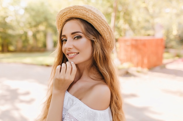 Close-up portret van fascinerende jonge vrouw speels poseren in zonnige dag staande in park. Buiten foto van prachtig meisje met mooie lichtgroene ogen glimlachen, haar kin aan te raken.