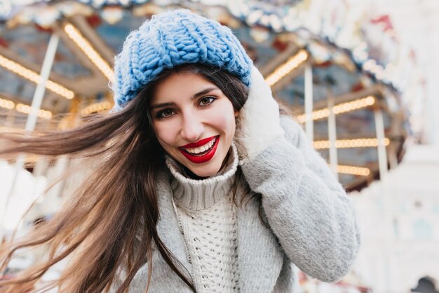 Close-up portret van geïnspireerd meisje in wollen blauwe hoed poseren met een glimlach op de achtergrond wazig. prachtige brunette dame in warme kleren die plezier heeft in het pretpark tijdens de kerstvakantie.