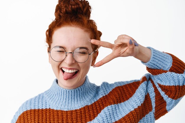 Close-up portret van gelukkig en positief roodharige meisje in glazen, tong met v-teken tonen en plezier hebben, vreugde en opwinding uitdrukken op wit