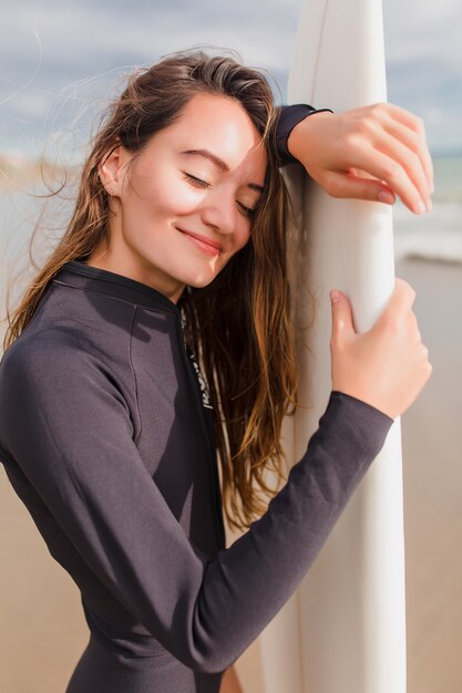 Close-up portret van gelukkig lachend mooi meisje met lang haar en charmante glimlach staat in het zonlicht aan de oever van de oceaan en kalmte bewaren. Actieve sportieve vrouw bij zonnig weer met surfplank