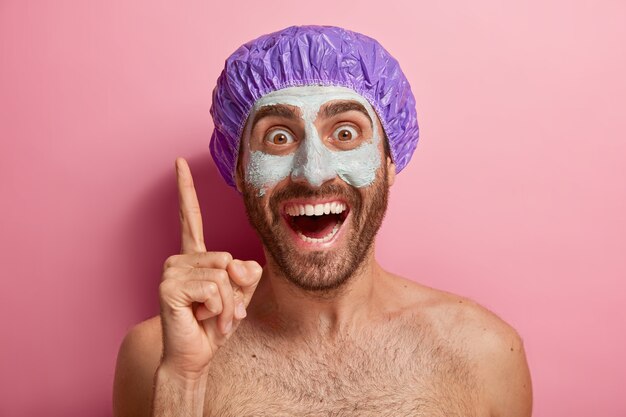 Close-up portret van gelukkig mannelijk model verhoogt wijsvinger, naar boven wijst, draagt kleimasker op gezicht