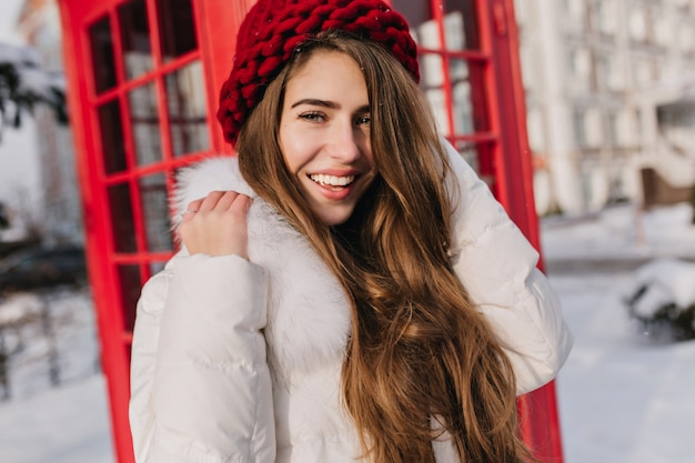 Close-up portret van gelukkige vrouw met glanzend bruin haar poseren naast rode telefooncel. Buitenfoto van een prachtig vrouwelijk model in gebreide baret die geniet van een ijzige ochtend in Engeland.