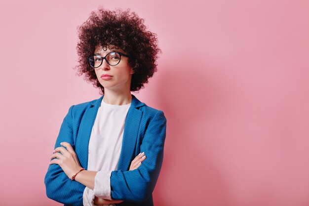 Close-up portret van helder gekrulde vrouw gekleed blauwe jas en bril staat met ernstige emoties op roze