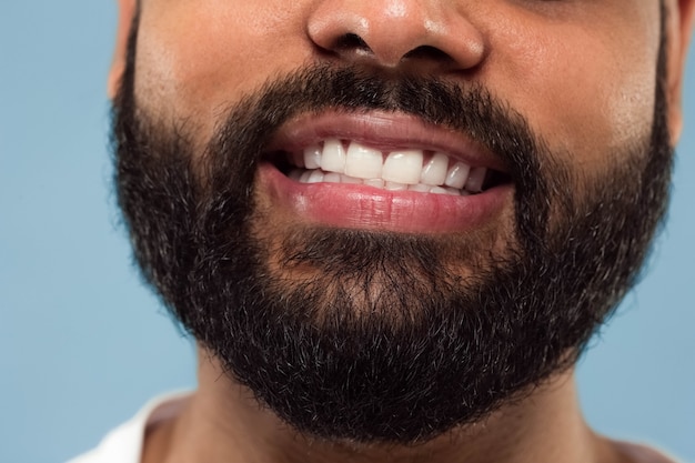 Close-up portret van het gezicht van de jonge hindoe man met baard, witte tanden en lippen op blauwe achtergrond. Lachend. Menselijke emoties, gezichtsuitdrukking, reclameconcept. Negatieve ruimte.