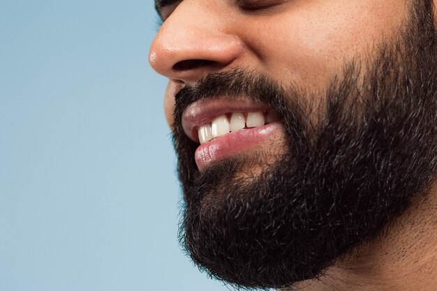 Close-up portret van het gezicht van de jonge hindoe man met baard, witte tanden en lippen op blauwe muur. Lachend. Menselijke emoties, gezichtsuitdrukking, reclameconcept. Negatieve ruimte.