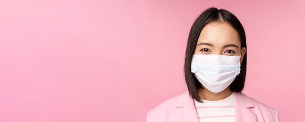 Close-up portret van japanse zakenvrouw in medisch gezichtsmasker pak kijkend naar camera staande over roze achtergrond