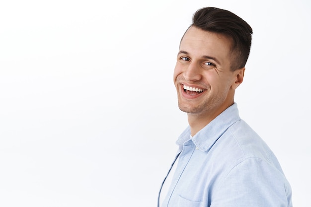 Close-up portret van knappe stijlvolle jongeman die in profiel staat, hoofd draait met een stralende glimlach, tevredenheid en enthousiasme uitdrukt, staande witte muur tevreden