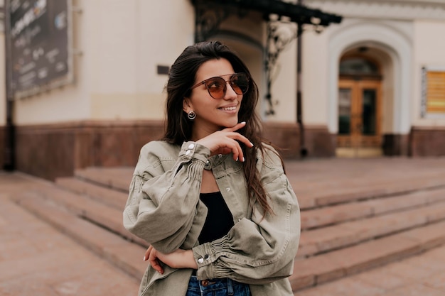 Close-up portret van lachen brunette meisje lopen op de achtergrond van de stad. Gelukkige vrouw in zonnebril en kleding die buiten lopen. Afbeelding van mooie stijlvolle vrouw op straat op lentedag