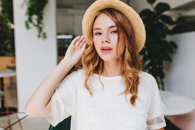 Close-up portret van licht gebruind meisje met elegante make-up poseren met hand omhoog in gezellige kamer. Binnenfoto van een schattige, krullende jonge dame met een bleke huid en een serieus gezicht op een wazige achtergrond.