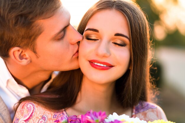 Close-up portret van mooi paar verliefd, knappe vriend zijn vrouw kussen op de wang, zonsondergang kleuren, lichte make-up, elegante kleren laksheid romantische datum.