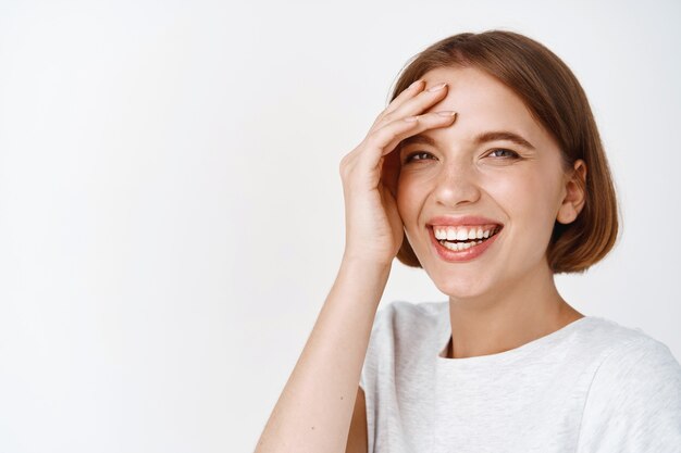 Close-up portret van mooi vrouwelijk model met kort haar, zorgeloos glimlachen en lachen, opgewonden gezicht aanraken, staande tegen een witte muur