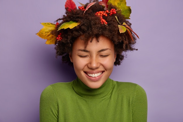 Close-up portret van mooie Afro-Amerikaanse vrouw lacht hardop, heeft brede brede glimlach, houdt de ogen gesloten van plezier, draagt herfstbladeren vast in haar in goed humeur