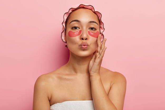 Close-up portret van mooie vrouw heeft natuurlijke schoonheid, houdt de lippen gevouwen om iemand te kussen, draagt een waterdichte badmuts, raakt de wang aan, geniet van de frisheid van de huid, heeft ooglapjes op het gezicht, poseert binnen