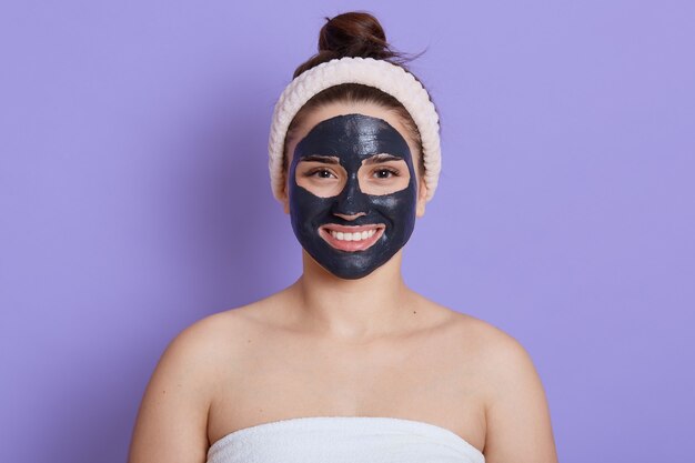 Close-up portret van mooie vrouw met zwart gezichtsmasker geïsoleerd over lila muur, meisje met witte handdoek op haar lichaam en blote schouders, tevreden en gelukkige glimlach, reinigingsprocedure aan het doen.