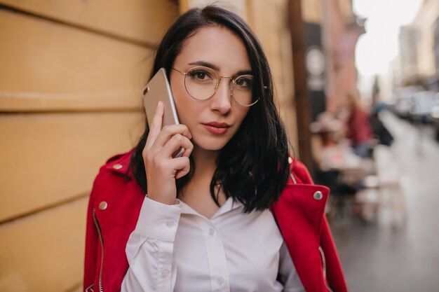 Close-up portret van mooie zwartharige vrouw in rode jas poseren met smartphone op stadsmuur