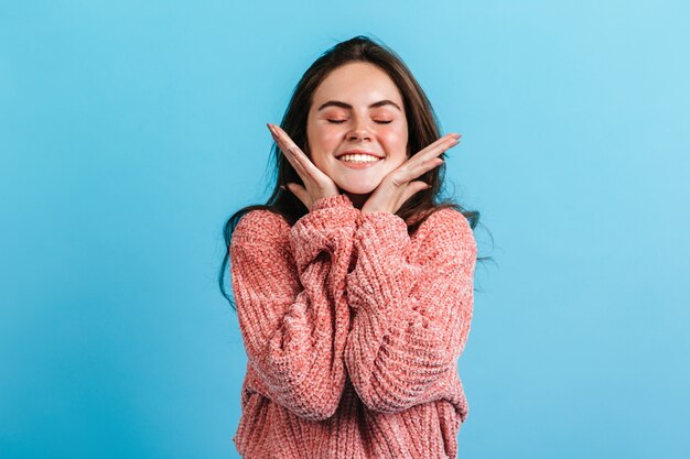 Close-up portret van schattige brunette lachend met gesloten ogen. Meisje in roze trui raakt zachtjes haar wangen.