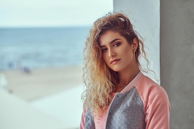 Close-up portret van sensueel jong meisje met krullend blond haar gekleed in een roze t-shirt en jas poseren tegen de zeekust.