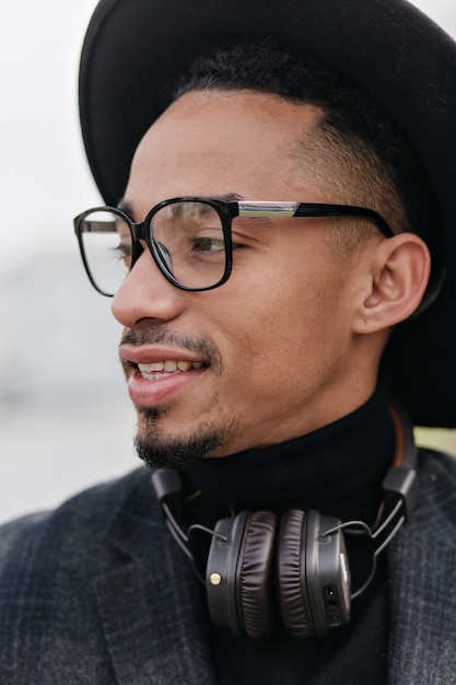 Close-up portret van verfijnde jonge man met bruine huid. Foto van dromerig Afrikaans mannelijk model in glazen en hoofdtelefoons die openlucht koelen.