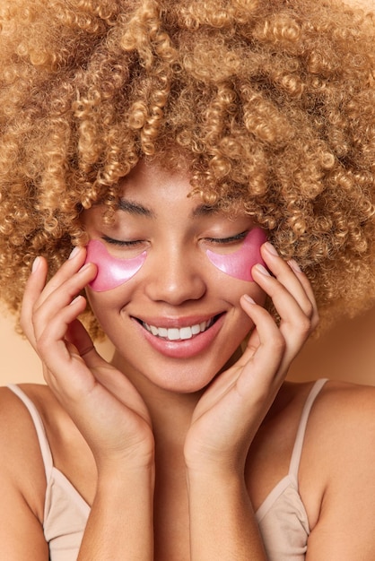 Close-up portret van vrolijke vrouw met krullend haar glimlacht graag hydrogel patches onder de ogen voor het verminderen van rimpels houdt de ogen gesloten geniet van schoonheidsbehandelingen. Huidverzorging en verjonging