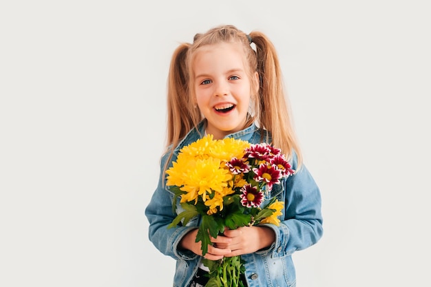 Close-up, selectieve aandacht. klein blond meisje in een houdt chrysanten en gerbera's in haar handen op een witte achtergrond, een kindmeisje glimlacht en houdt lentebloemen in haar handen