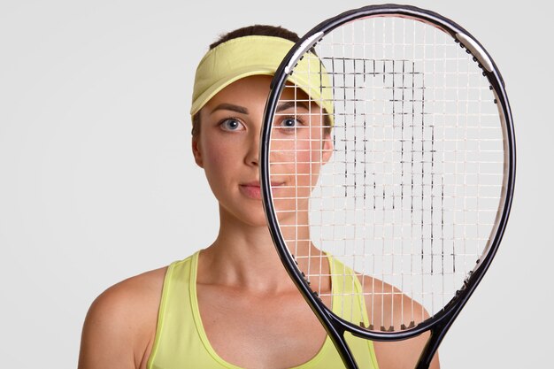 Close-up shot van aangenaam ogende gezonde vrouw houdt tennisracket, wordt runner up, kijkt door net, draagt court cap