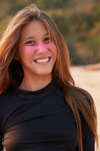 Close-up shot van aantrekkelijke surfgirl gekleed in zwart wetsuit, heeft roze masker op haar mooie gezicht, heeft lang haar