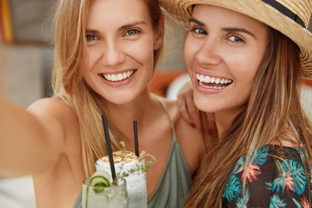 Close-up shot van blonde en brunette vrouwen hebben een brede glimlach, poseren voor de camera en maken selfie, houden exotische cocktails, hebben zomervakanties. Mensen, geluk, recreatie en levensstijlconcept