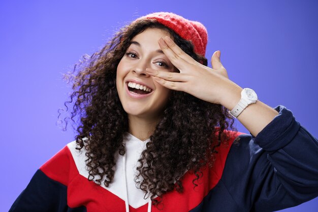 Close-up shot van charismatische gelukkig zorgeloze vrouw in winter rode schattige muts en sweatshirt met vingers in de buurt van oog en gluren met brede glimlach op camera met plezier, spelen op blauwe achtergrond.