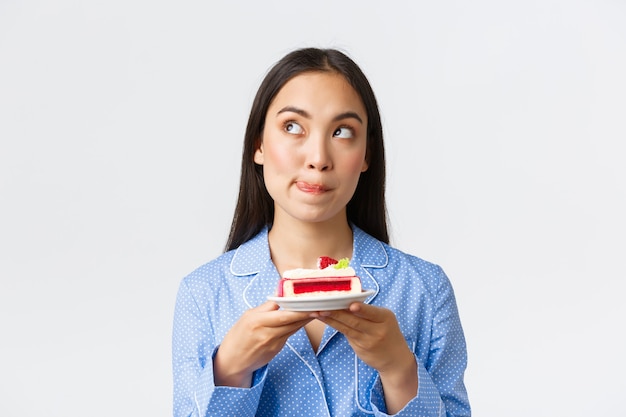 Close-up shot van doordacht schattig aziatisch meisje dat 's nachts in pyjama staat met een bord cake, op zoek naar de linkerbovenhoek beslissend, kan de verleiding niet weerstaan, eet heerlijk dessert, witte achtergrond