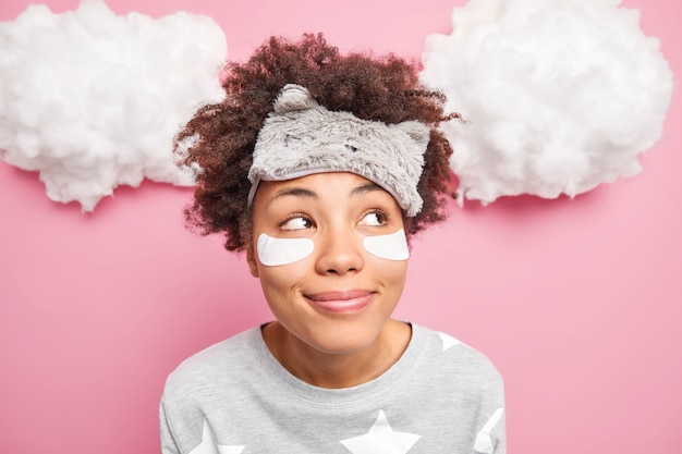 Close-up shot van dromerige schattige krullende vrouw kijkt peinzend opzij denkt aan iets aangenaams draagt slaapmasker op voorhoofd nachtkleding geniet van goedemorgen en begin van de nieuwe dag