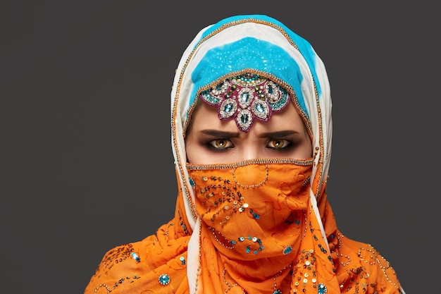 Close-up shot van een mooie jonge vrouw met professionele make-up, gekleed in een elegante kleurrijke hijab versierd met pailletten en sieraden. Ze poseert in de studio en kijkt boos op een donkere achtergrond