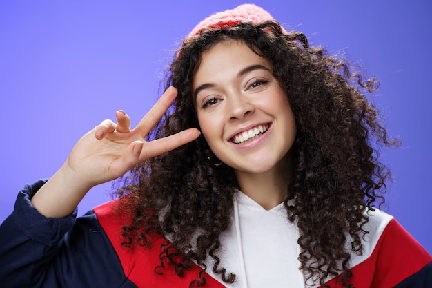 Close-up shot van een vriendelijke charmante vrouw met krullend haar met een warme beanie die het hoofd kantelt en vreugdevol glimlacht en een overwinnings- of vredesgebaar in de buurt van het gezicht laat zien, zich gelukkig en opgetogen over de blauwe muur.